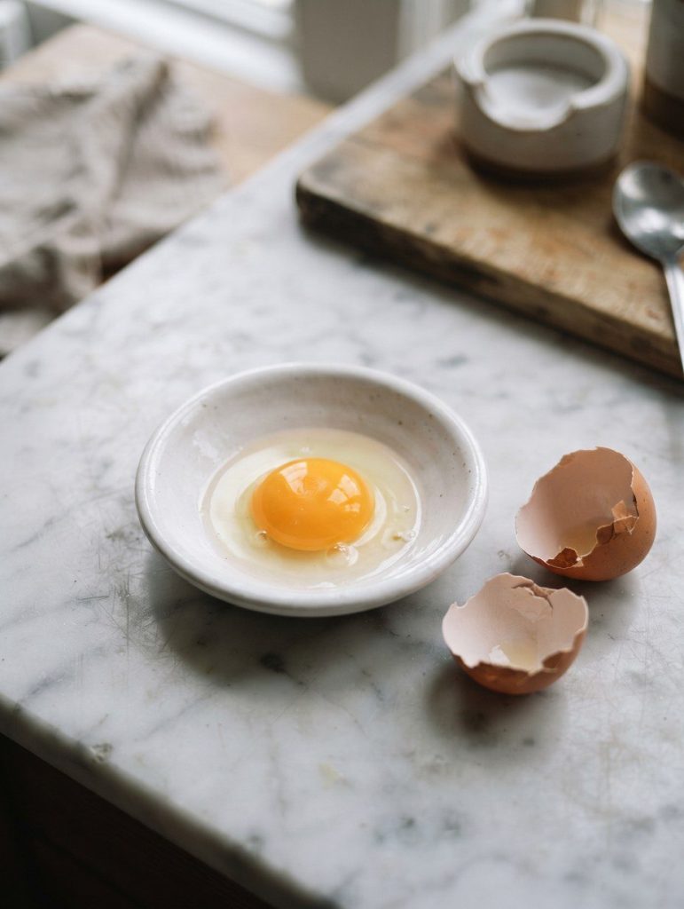 A raw egg yolk in a shallow white dish beside a cracked shell