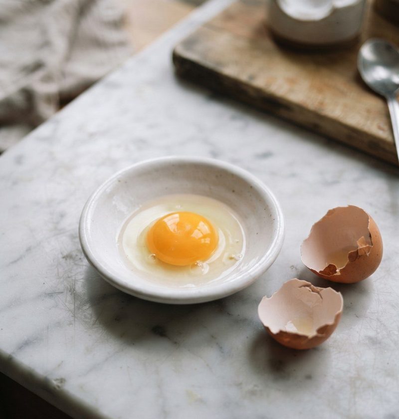 A raw egg yolk in a shallow white dish beside a cracked shell
